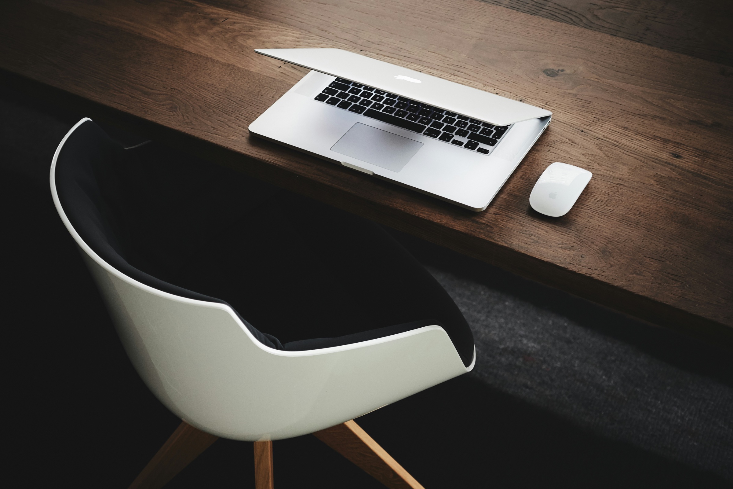 A laptop and mouse on a wooden desk beside a modern chair.